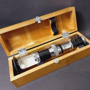 Könnte beinhalten: Ein Vintage-Mikroskop in einer offenen Holzkiste. Das Mikroskop hat einen schwarzen und silbernen Körper mit einer Linse an einem Ende. Die Kiste ist aus hellem Holz gefertigt und hat Metallscharniere und -verschlüsse.