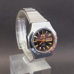 May include: A silver-toned Orient watch with a black face and gold accents. The watch features a day and date display and a silver link band. The watch is displayed on a clear stand against a dark background.