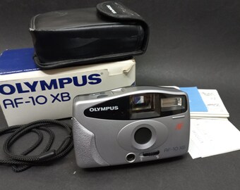 May include: A silver Olympus AF-10 XB point and shoot camera with a black strap and a black carrying case. The camera has a built-in flash and a red label that says "AF-10 XB".