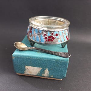 May include: A vintage salt cellar with a silver-colored metal base and a clear glass bowl. The base is decorated with blue and red enamel floral designs. A small silver spoon rests beside the salt cellar on a blue box.