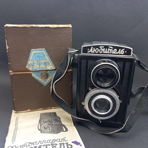 Puede incluir: Una cámara de película vintage negra y plateada con una correa de cuero y una caja marrón. La cámara tiene el texto "Любитель 16" en la parte delantera.