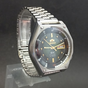 Könnte beinhalten: Eine silberne Orient SK Crystal 21 Jewels Armbanduhr mit schwarzem Zifferblatt und silbernem Metallband. Das Zifferblatt der Uhr hat ein Datumsfenster mit der Aufschrift "SON 29".