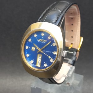 Könnte beinhalten: Eine goldfarbene Loreena Automatic Uhr mit blauem Zifferblatt und diamantartigen Akzenten. Die Uhr hat ein schwarzes Lederarmband und ein Datumsfenster.
