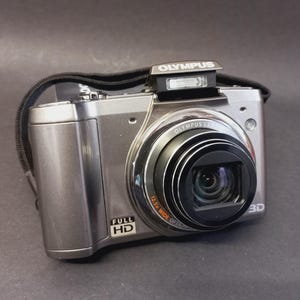 May include: Silver Olympus digital camera with a black strap. The camera has a prominent lens with the text "12.5x Wide Optical Zoom" and "FULL HD" printed on the body. The Olympus logo is visible on the top.