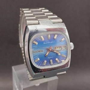 Puede incluir: Un reloj de pulsera vintage plateado con una esfera azul cuadrada. La esfera del reloj muestra la palabra "automatic" y la fecha. La correa del reloj está hecha de eslabones de metal plateado. El reloj tiene una corona y agujas plateadas.