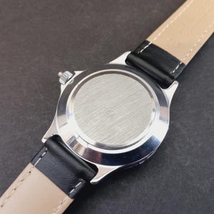 May include: Close-up of a silver-toned wristwatch with a black leather band and a beige underside. The watch face is not visible. The watch band is attached to the watch case with a silver-toned metal. The watch is on a dark surface.