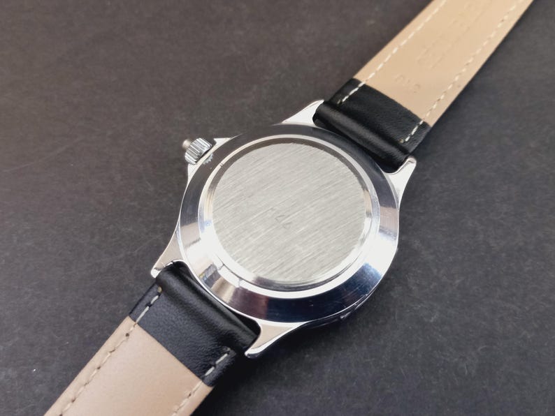 May include: Close-up of a silver-toned wristwatch with a black leather band and a beige underside. The watch face is not visible. The watch band is attached to the watch case with a silver-toned metal. The watch is on a dark surface.