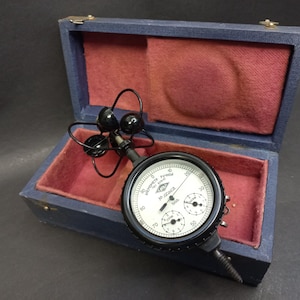 Puede incluir: Un medidor antiguo en blanco y negro con un dial y varios diales más pequeños, alojado en una caja de madera azul con un interior de terciopelo rojo. El medidor tiene un mango negro y un diseño complejo.