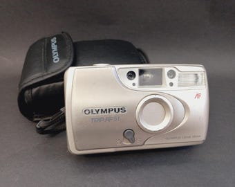 May include: A silver Olympus Trip AF 51 point-and-shoot camera with a 28mm lens, featuring an autofocus system, a built-in flash, and a viewfinder. The camera is accompanied by a black protective carrying case with the Olympus brand name printed on it.