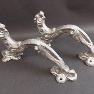 May include: Two silver-colored metal door handles featuring an ornate, curved design with sculpted details at the ends and mounting plates with pre-drilled holes for installation. The handles have a textured, matte finish and a distinct arched shape.