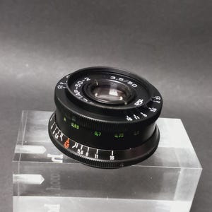 Op de afbeelding: Zwarte cameralens met de tekst "3.5/50" en "IAP-50-2". De lens heeft een geribbelde focusring en diverse meetmarkeringen. De lens rust op een helder, rechthoekig blok.