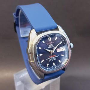 May include: A Seiko 5 Superior automatic wristwatch with a dark blue dial, silver-tone hands, and hour markers. The watch features a titanium case, a day-date window at the three o'clock position, and a smooth blue silicone strap.