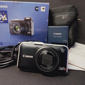 May include: A black Canon PowerShot SX230 HS digital camera with a 14x optical zoom lens, accompanied by its original blue product box, a black Acropolis carrying case, a battery charger, a black power cable, and a printed instruction manual.
