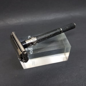 May include: A black and silver safety razor with a textured handle rests on a clear acrylic block. The razor features a silver head and a black, patterned handle with silver accents. The background is a dark gray.
