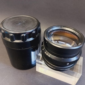 Op de afbeelding: Een zwarte cameralens met een zilveren ring en een zwarte opbergkoffer. De lens heeft een reeks nummers en markeringen op de zilveren ring. De opbergkoffer is cilindrisch met een geribbelde bovenkant.