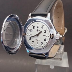 May include: A silver-toned wristwatch with a white face, black numerals, and hands. The watch has a hinged cover and a black leather band. The watch face displays the word "Luch" and "Quartz".