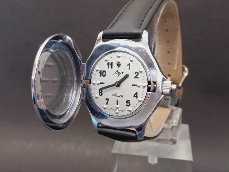 May include: A silver-toned wristwatch with a white face, black numerals, and hands. The watch has a hinged cover and a black leather band. The watch face displays the word "Luch" and "Quartz".