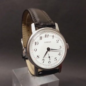 Puede incluir: Un reloj de pulsera vintage con esfera blanca, manecillas negras y la palabra "ПОБЕДА" impresa en la esfera. El reloj tiene una correa de cuero negro y una caja plateada. El reloj se exhibe sobre un soporte transparente sobre un fondo oscuro.