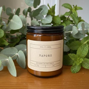 May include: An amber glass jar candle with a black screw-top lid and a cream-colored label. The label features the text SOL Y CERA, VAPORU, ARTISANAL SOY WAX CANDLE, 8 oz / 227g, and 40hr. The candle is positioned in front of green eucalyptus leaves.