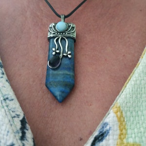 Puede incluir: Un collar con un colgante de plata adornado con una piedra preciosa azul y verde. El colgante tiene forma de lágrima y está decorado con un diseño de filigrana plateado.