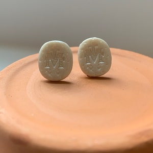 Puede incluir: Un par de pendientes ovalados blancos con un diseño en relieve de una cruz y las letras "M" y "A".