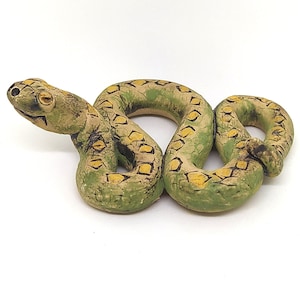 May include: A ceramic snake figurine in shades of green, tan, and yellow. The snake is coiled, with a detailed pattern of yellow diamond shapes on its back. The figurine has a textured appearance and is set against a white background.
