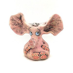 May include: A pink ceramic elephant figurine with large ears, blue eyes, and orange feet. The elephant is sitting with its trunk raised.
