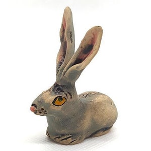 May include: A small ceramic rabbit figurine with grey and brown fur, painted with a yellow eye and a pink nose. The rabbit is sitting with its ears up.