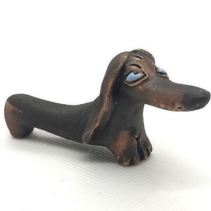 May include: A small, brown ceramic dachshund figurine with blue eyes. The figurine is hand-painted and has a whimsical expression.