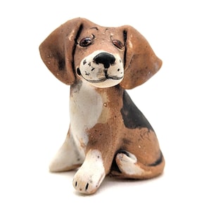 May include: A ceramic beagle figurine in a seated position. The dog has brown and white fur with black markings. The figurine has large, floppy ears and a detailed face with dark eyes and a black nose. The overall design is rustic and handcrafted.