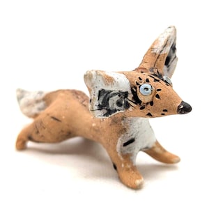 Peut inclure: Petite figurine de renard en céramique faite à la main. Le renard a un corps beige avec des marques blanches et noires sur les oreilles et la queue. Il a des yeux bleus et une surface texturée détaillée. La figurine est posée dans une posture ludique.