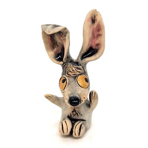 May include: A small ceramic figurine of a creature with large, upright ears, oversized yellow eyes, and a black nose. The figure has a light grey body with dark grey accents, a tuft of hair on its head, and two small arms raised in a waving gesture.