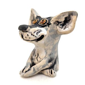 May include: A ceramic dog figurine with a playful design. The dog features large ears, a black nose, and expressive eyes. The figurine is painted in shades of grey, black, and beige, with a smiling expression. The dog is in a seated position.