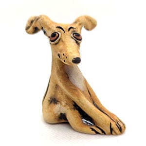 May include: A whimsical ceramic figurine of a greyhound dog in a seated position. The dog has a tan body with black spots, large brown eyes, and a black nose. The ears are perked up, and the overall expression is friendly.