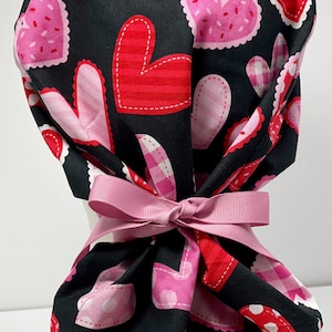 May include: A black scrub hat with a pink ribbon tie. The hat is covered in a pattern of pink, red, and white hearts.