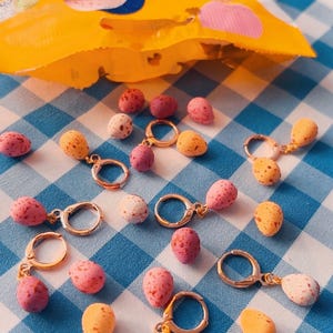 May include: Speckled egg-shaped earrings in various colours, including pink, yellow, and white, with gold-coloured hoops. The earrings are displayed on a blue and white gingham surface, with a yellow bag visible.