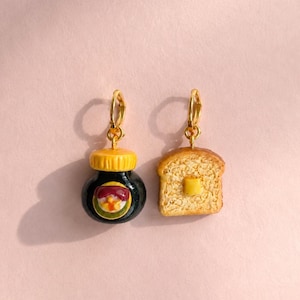 May include: Two gold-toned metal charms featuring food-themed designs. One charm is a black jar with a yellow lid and a small label, and the other is a slice of toasted bread with a yellow square of butter on top. Both charms have gold hoops.