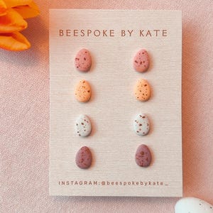 May include: Eight speckled egg-shaped earrings in pink, orange, white, and purple are displayed on a beige card. The card reads "BEESPOKE BY KATE" and "INSTAGRAM:@beespokebykate_". Orange and yellow tulips are in the background.
