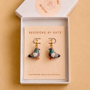 May include: A pair of gold hoop earrings featuring small, hand-painted pigeon charms in shades of grey, green, and white with orange feet. The earrings are displayed on a branded card inside a white gift box labelled BEESPOKE BY KATE.