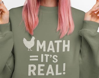 Może przedstawiać: Oliwkowa bluza z napisem "MATH = IT'S REAL!" i grafiką kurczaka. Osoba na zdjęciu ma na sobie niebieską czapkę i różowe włosy.