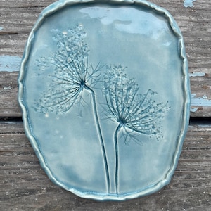 May include: A light blue ceramic plate with a raised floral design. The oval-shaped plate has a textured, scalloped edge and features an embossed image of two stylized flowers. The plate is displayed on a weathered wooden surface.