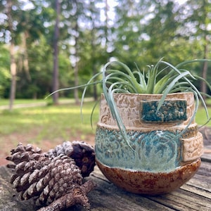 May include: A small, round, handmade ceramic planter with a textured, multi-colored glaze in shades of tan, teal, and dark brown. The pot features raised rectangular accents and contains a spiky, green air plant. Two pinecones sit nearby.