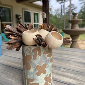 May include: A ceramic vase with a floral pattern in brown and blue-green tones. The vase holds several beige, spherical objects and dried seed pods. The arrangement sits on a wooden surface, with a blurred outdoor background.