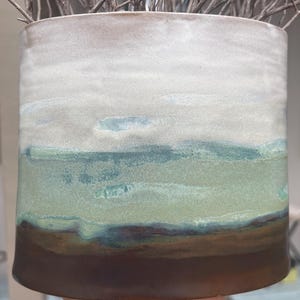 May include: A cylindrical ceramic planter featuring a horizontal gradient glaze. The top section is a matte off-white, transitioning into a soft sage green middle band, and finishing with a solid dark brown base. Bare twigs extend from the top.