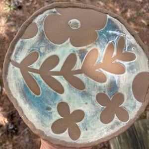 Puede incluir: Un plato de cerámica hecho a mano con un borde marrón y un diseño floral. El plato presenta un esmalte azul claro y blanco con flores y hojas recortadas en marrón. El plato es redondo y tiene un aspecto rústico y orgánico.