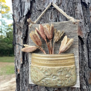 May include: A wall-mounted planter featuring a rectangular weathered wooden board with a jute twine hanger. Attached to the board is a semi-circular ceramic pocket in a muted olive green glaze with a raised floral relief pattern, holding dried seed pods.