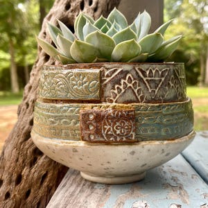May include: A ceramic planter with a light green succulent plant. The pot features a speckled white base and two textured, layered bands in shades of blue, green, and brown, decorated with intricate floral and geometric embossed patterns.
