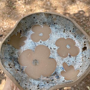 May include: A handmade ceramic bowl with an irregular shape. The bowl has a speckled, blue-grey glaze and brown flower cutouts. The rim is a darker brown, giving a rustic and artistic feel.