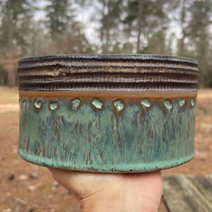 May include: A handcrafted ceramic bowl with a textured, dark brown rim and a turquoise-green body. The bowl features a band of brown with small, circular accents. The exterior has a rustic, glazed finish, perfect for serving or display.