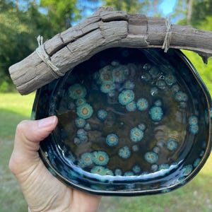 May include: A round ceramic bowl with a dark brown and black glossy glaze featuring turquoise and gold crystalline spots. A piece of weathered driftwood is attached to the top rim of the bowl with thin, light-colored twine wrapped around it.